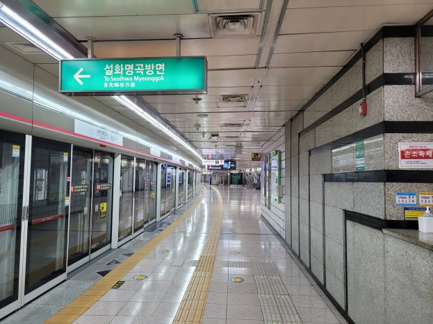 Ein belebter Seoul-U-Bahnhof mit einem Zug auf der linken Seite und einem Bahnsteig auf der rechten Seite mit an der Wand montierten Tafeln und einem grünen Deckenzeichen, während Menschen auf den Zug warten.