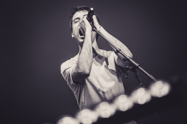 Ein Mann singt in ein Mikrofon auf einer hell erleuchteten Bühne, trägt ein schwarzes T-Shirt und Jeans, hat verwuschelte Haare und geschlossene Augen.