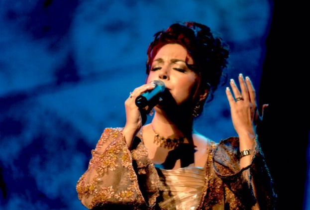 Eine Frau in einem goldenen Kleid singt in ein Mikrofon vor einem blauen und schwarzen Hintergrund, mit ESC-Text unten.