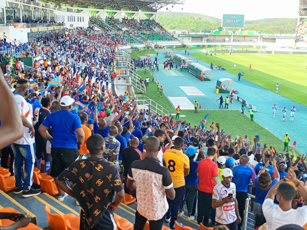 Eine große Menschenmenge versammelt sich vor einem eingezäunten Fußballfeld, einige sitzen auf Stühlen, andere stehen, unter einem klaren blauen Himmel mit Hügeln im Hintergrund und warten auf den Beginn des AFCON 2019-Spiels.