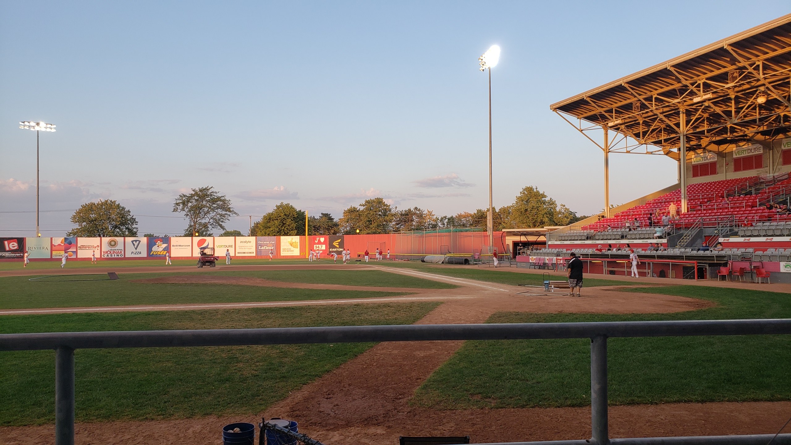 Baseball-Spiel im Gange mit Zuschauern in der Nähe eines Zaunes, umgeben von Stadion-Infrastruktur und einem bewölkten Himmel.