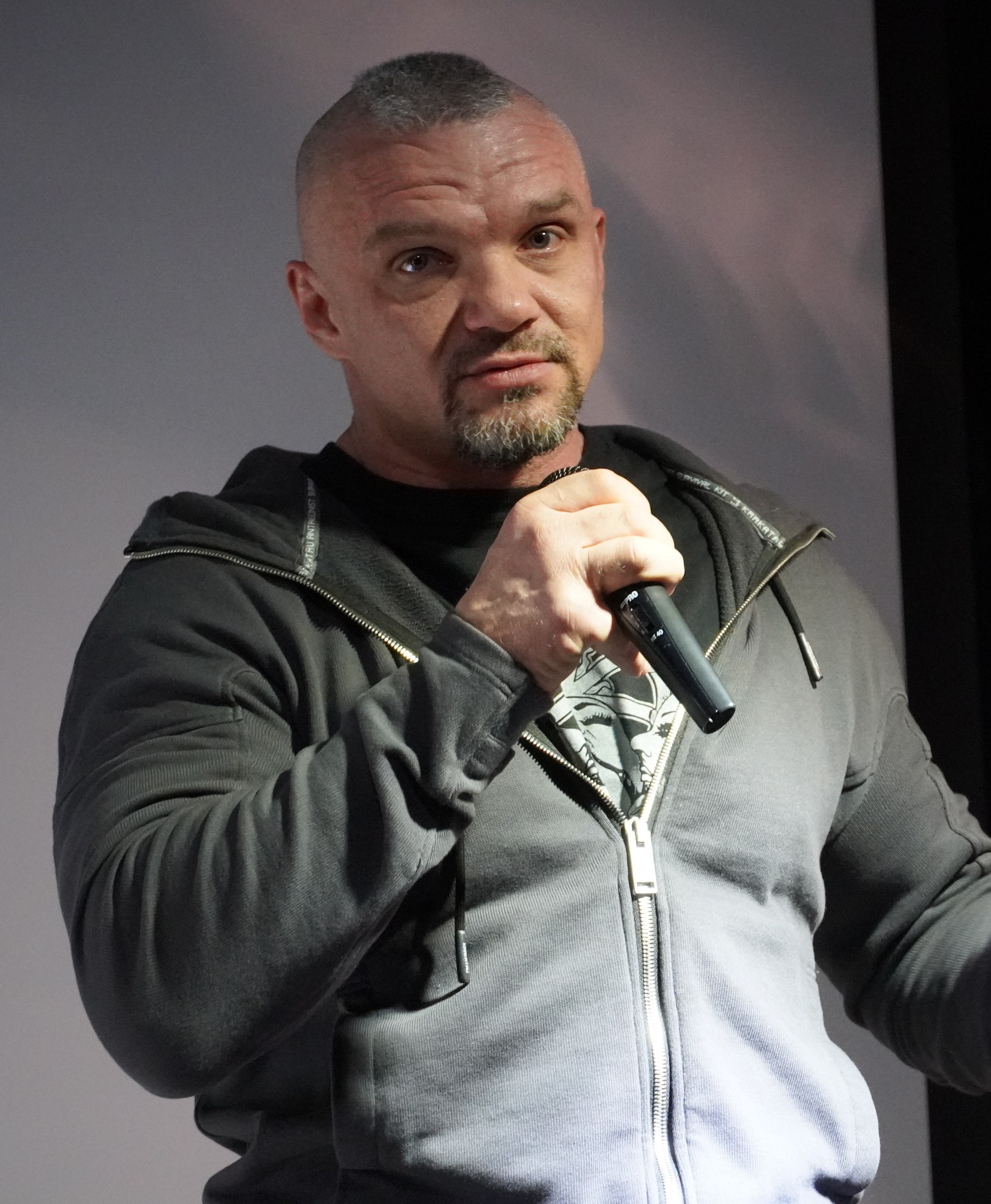 Ein Mann in einer schwarzen Kapuzenjacke und einem schwarzen Anzug mit einem weißen Hemd, der ein Mikrofon hält, steht selbstbewusst vor einer Wand und sieht aus wie ein Comedian mit zurückgekämmtem Haar.