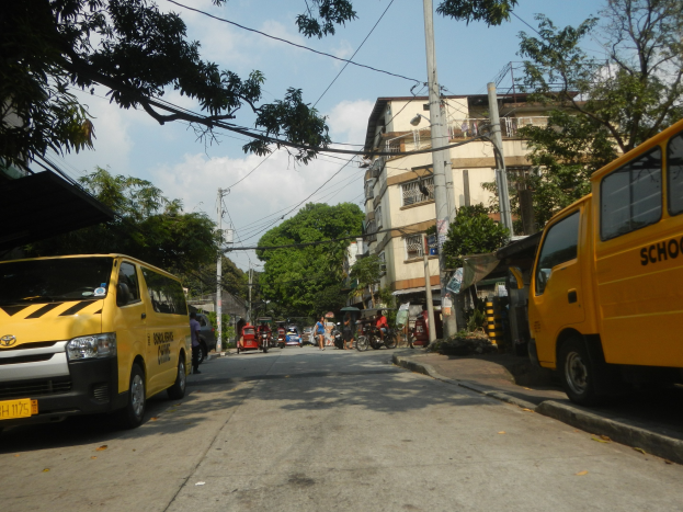 Ein gelber Schulbus fährt eine Straße mit hohen Gebäuden, Bäumen und Strommasten mit Drähten entlang, mit Menschen auf Fahrrädern und anderen Fahrzeugen auf der Straße, unter einem bewölkten Himmel.