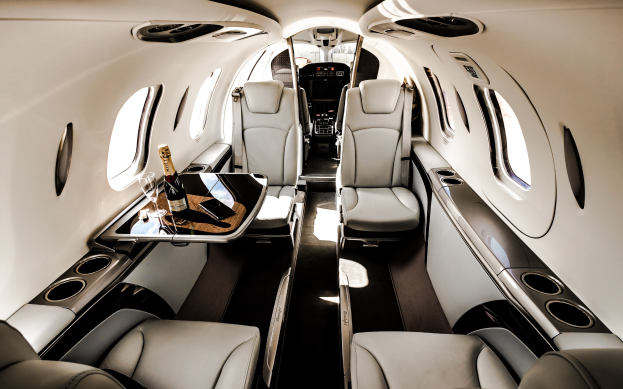 Das Innere eines Privatjets mit einem zentralen Tisch, auf dem sich zwei Flaschen und zwei Gläser befinden, sowie luxuriösen Polstern und Fenstern, die den Blick nach draußen ermöglichen.