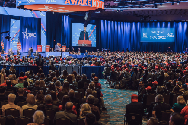 Eine große Menschenmenge sitzt in Reihen vor einer Bühne mit Personen auf dem Podium und links davon, blaue Vorhänge und ein Bildschirm im Hintergrund und ein orangefarbenes Schild oben mit der Aufschrift "CPAC 2020 Oktober 25."