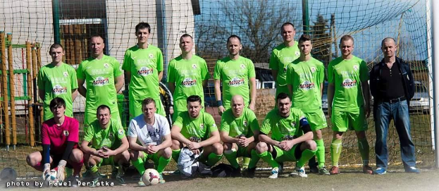 Eine Gruppe von Männern in grünen T-Shirts steht auf einem Fußballfeld, einige hocken, mit einem Ball davor und einem Torpfosten rechts daneben, vor einem Hintergrund aus Bäumen, Gebäuden und Fahrzeugen.