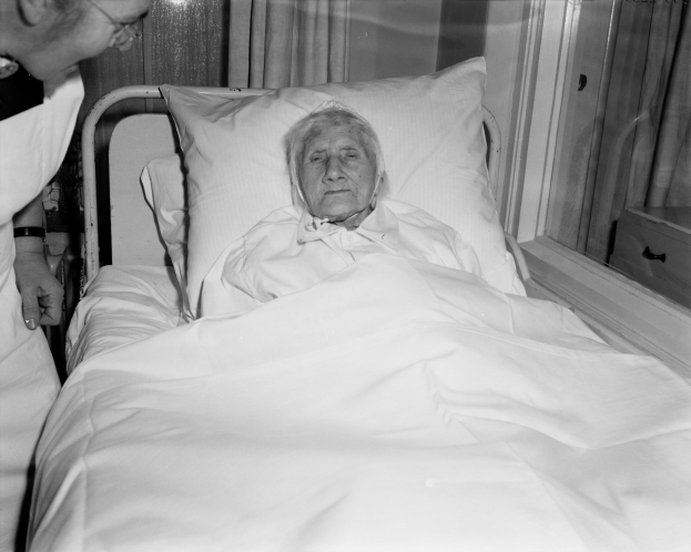 Ein Bild einer älteren Frau, die in einem Krankenhausbett liegt, mit einer Schwester neben ihr, das Bett mit einem weißen Laken und einem Kissen bedeckt und ein Fenster mit Vorhängen im Hintergrund.