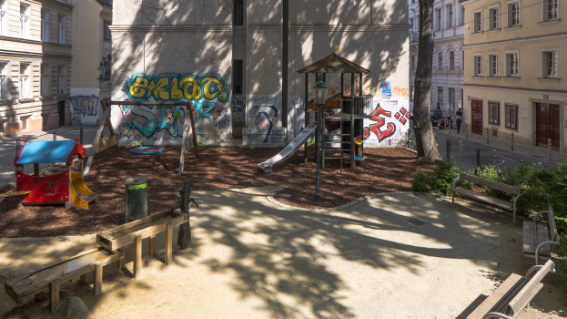 Spielplatz mit Graffiti, umgeben von Stadtgebäuden, Bäumen, Bänken und einigen Menschen und Fahrzeugen auf der Straße unter einem sichtbaren Himmel.