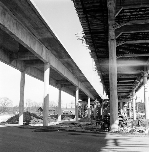 Schwarzes und weißes Foto einer Brücke im Bau, gestützt von Säulen auf der rechten Seite, mit einer Straße darunter, Bäumen und Häusern im Hintergrund und einem klaren Himmel darüber.