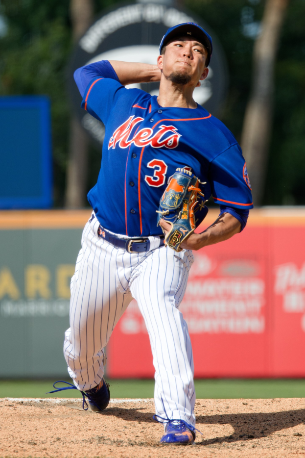 Ein Baseballspieler in einem blauen T-Shirt, weißen Hosen, blauer Mütze und blauen Schuhen wirft einen Ball auf einem Feld mit einer Werbetafel und Bäumen im Hintergrund.