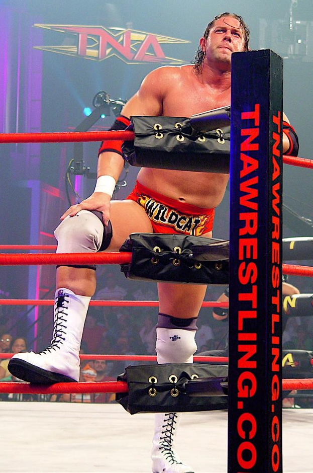 Ein Mann steht in einem Wrestling-Ring, die Hände auf den Knien, umgeben von Seilen und einer Stange mit Text, mit einer Menge und einem beleuchteten Schild im Hintergrund.