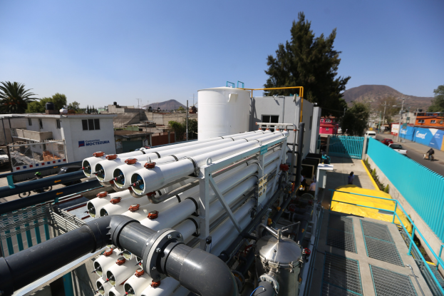 Große Gruppe von Industrieleitungen und Maschinen auf dem Dach eines Gebäudes mit Fahrzeugen, Menschen und Gebäuden in einer städtischen Umgebung unter einem klaren blauen Himmel.
