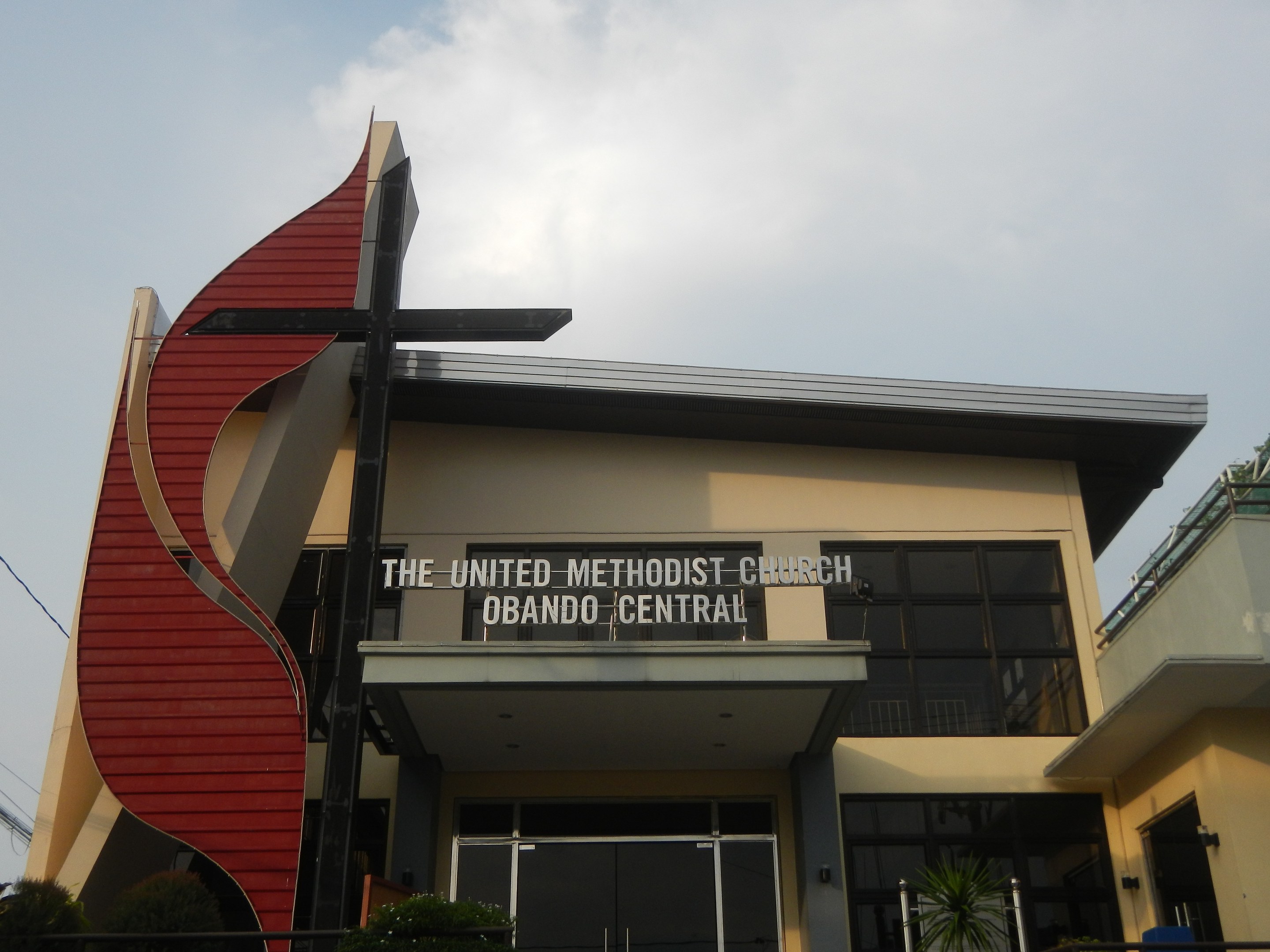 Außenansicht der United Methodist Church in Orlando, Florida, mit Glasfenstern, einer Namensplakette, Pflanzen, einem Metallzaun, Bäumen, Drähten und einem bewölkten Himmel.