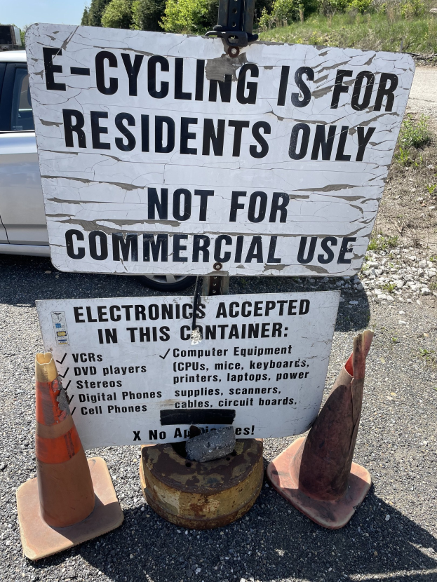 Sign reading "e-cycling is for residents only" placed beside a road, flanked by traffic cones and other objects, with a vehicle, trees, and a clear blue sky in the background.