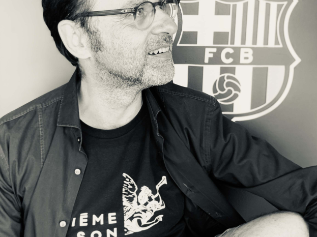 Schwarzes und weißes Foto eines grinsenden Mannes mit Brille und schwarzem Hemd, der vor dem FC Barcelona-Logo sitzt.