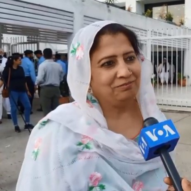 Eine Frau in einem weißen Sari mit einem Mikrofon, umgeben von einer Gruppe von Menschen im Freien mit einem Metallzaun, Topfpflanzen, einem Gebäude, einem Baum und dem Himmel im Hintergrund.