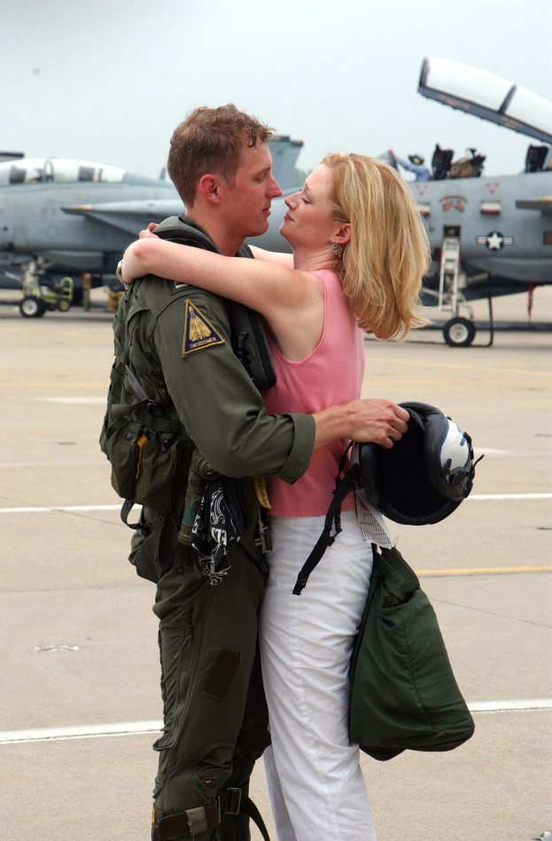 Ein Mann in einer grünen Jacke und weißen Hosen umarmt eine Frau mit einem schwarzen Helm und einer grünen Tasche vor Kampfjets unter einem klaren blauen Himmel.