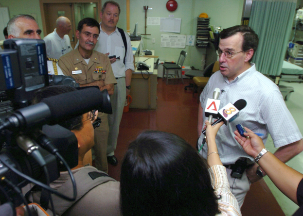 Eine Gruppe von Menschen mit Mikrofonen und Kameras umgibt einen Mann in einem weißen Hemd in einem Krankenhauszimmer mit Möbeln und medizinischen Geräten.