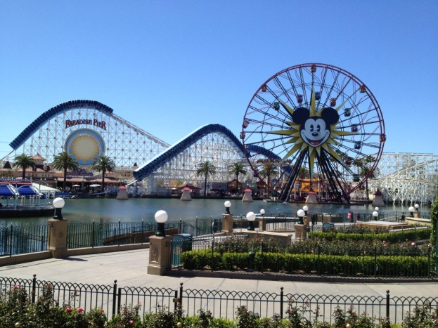 Der Disneyland California Adventure Freizeitpark mit einem Riesenrad und einer Achterbahn, umgeben von Grünflächen, Laternenmasten, einem Wasserbecken im Vordergrund und einem klaren blauen Himmel.
