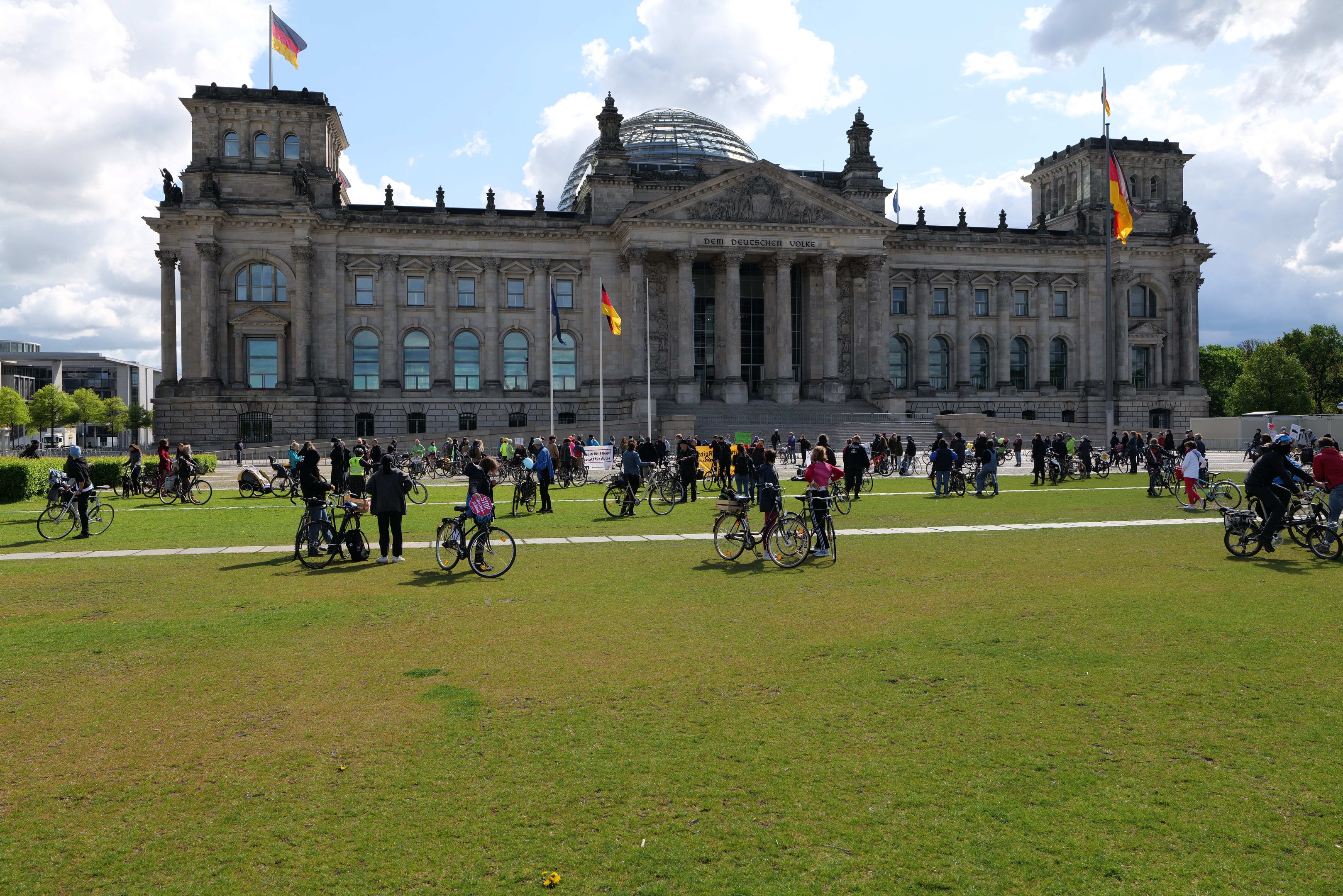 Eine Gruppe von Menschen, die vor dem Reichstaggebäude in Berlin, Deutschland, Fahrrad fährt, mit sichtbaren Fenstern, Säulen und Fahnenmasten, umgeben von Grün unter einem bewölkten Himmel.