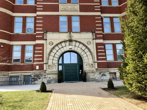 Großes Backsteingebäude mit einer grünen Tür und Fenstern, als Middle Park Public School identifiziert, umgeben von Pflanzen, Gras, einem Pfad, einem Metallzaun und einer Baumgruppe unter einem bewölkten Himmel.