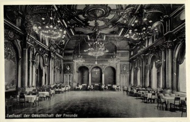 Black and white photo of a large room with tables, chairs, pillars, arches, and chandeliers, labeled "Festival der Gesellschaft der Freunde, Berlin" at the bottom.