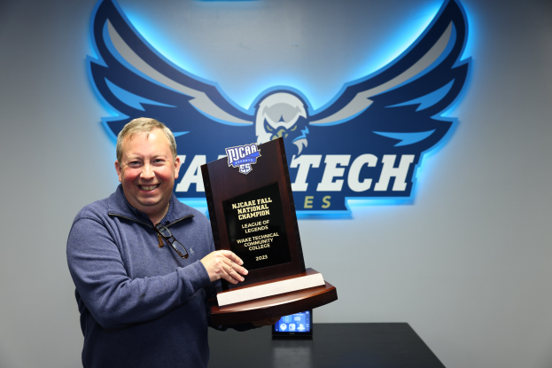 Ein Mann steht lächelnd vor einer Wand mit einem Logo und Text, während im Hintergrund ein Handy auf einem Tisch liegt.