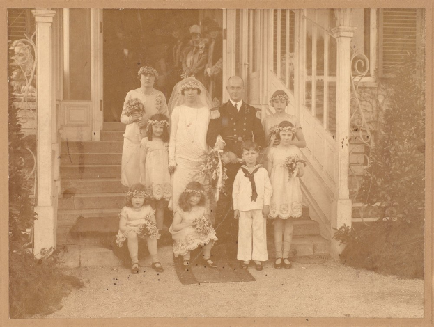 Altes Schwarz-Weiß-Foto einer Familie, die vor einem Haus posiert, mit Erwachsenen, die stehen, und Kindern, die auf den Stufen sitzen, jeder hält einen Blumenstrauß; Topfpflanzen flankieren die Szene und der Hintergrund zeigt Säulen, Fenster und eine Tür.