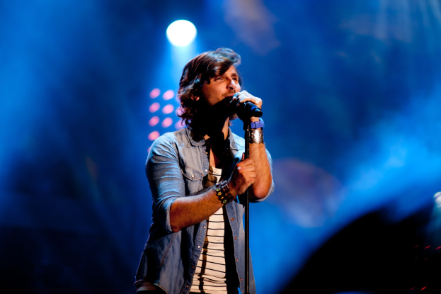 Ein Mann auf einer hell erleuchteten Bühne singt in ein Mikrofon und trägt ein schwarzes T-Shirt, Jeans und eine Gitarre, mit unordentlichem Haar und einem selbstbewussten Ausdruck.