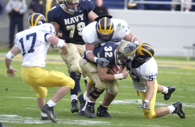 A football player in a helmet and uniform is being tackled by multiple defenders on a grassy field, with spectators and a wall visible in the background.