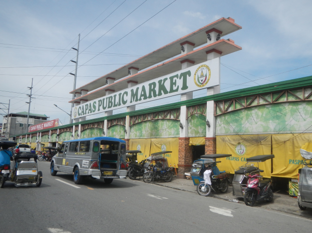 Bustling city street with cars, motorcycles, and rickshaws in front of "Capas Public Market" building, electric poles, light poles, and cloudy sky in the background.