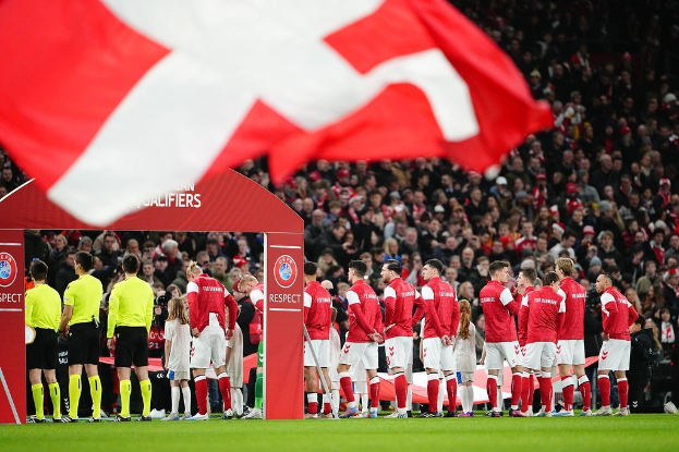 Gruppe von Menschen auf einem Fußballfeld mit einer roten und weißen Flagge im Vordergrund, einem Bogen mit der Aufschrift "Bayern Munich vs Bayern Munich Betting Tips & Preview" im Hintergrund und einem großen Stadionpublikum.