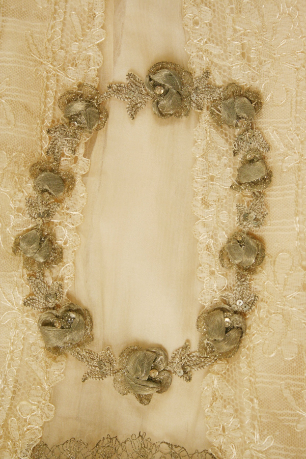 Nahaufnahme eines weißen Kleides mit aufwendiger Spitzenverzierung und einer goldenen Kette darauf.