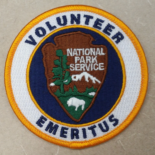 Runder Patch mit blauem Rand und weißem Hintergrund, der das zentrierte schwarze Text "Volunteer National Park Service Emeritus" in einem gelben Kreis enthält.