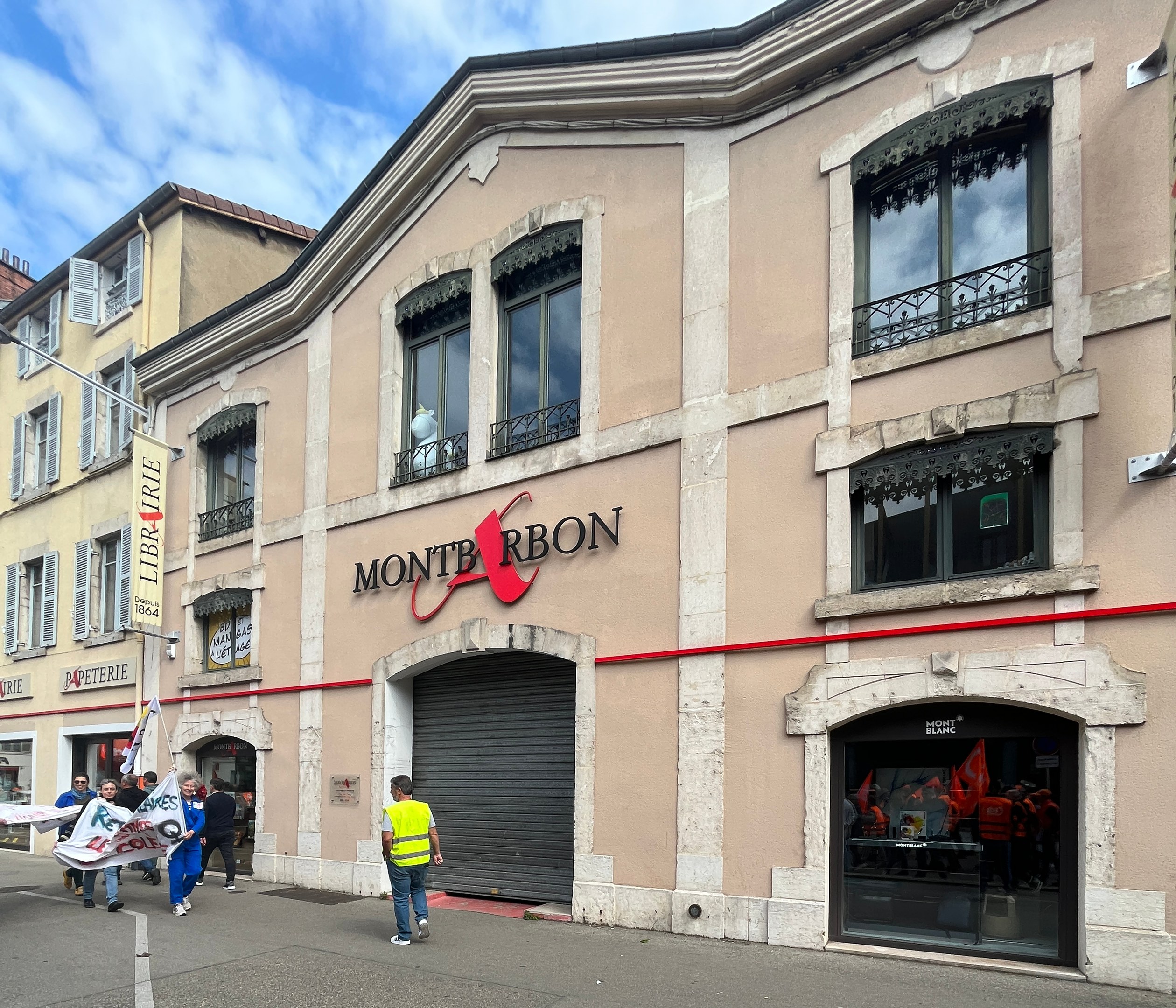 Eine Gruppe von Menschen geht eine Straße entlang vor einem Gebäude mit einer 'Montbron'-Schildertafel, einem Straßentisch, einer Fahne und einem bewölkten Himmel, während sie Fahnen schwenken.