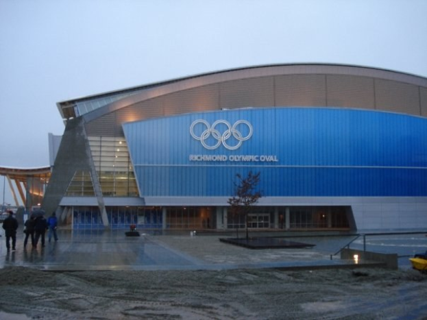 Olympiastadion in Sotschi, Russland, mit Füülern, einem geparkten Fahrzeug, Bäumen, Gebäuden und einem klaren blauen Himmel im Hintergrund.