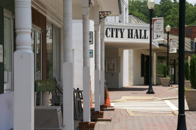 Rathausgebäude mit umgebender Infrastruktur, einschließlich Straßenlaternen, Schildern, Fahrzeugen, Verkehrskegeln, Stühlen, Topfpflanzen, Bäumen und einem bewölkten Himmel.