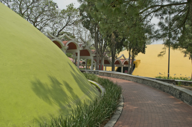 Ein Weg in einem Park mit einer grünen Wand auf der linken Seite, umgeben von Pflanzen und Bäumen, mit Gebäuden und einem klaren blauen Himmel im Hintergrund.