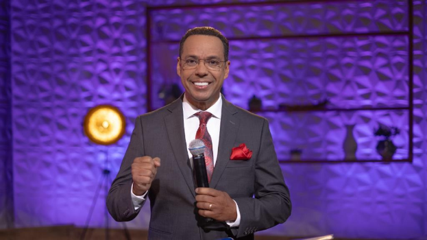 Stephen Colbert, host of "The Daily Show with Trevor Noah," stands on a stage holding a microphone, wearing a suit, tie, and spectacles, with a light and stand visible behind him.