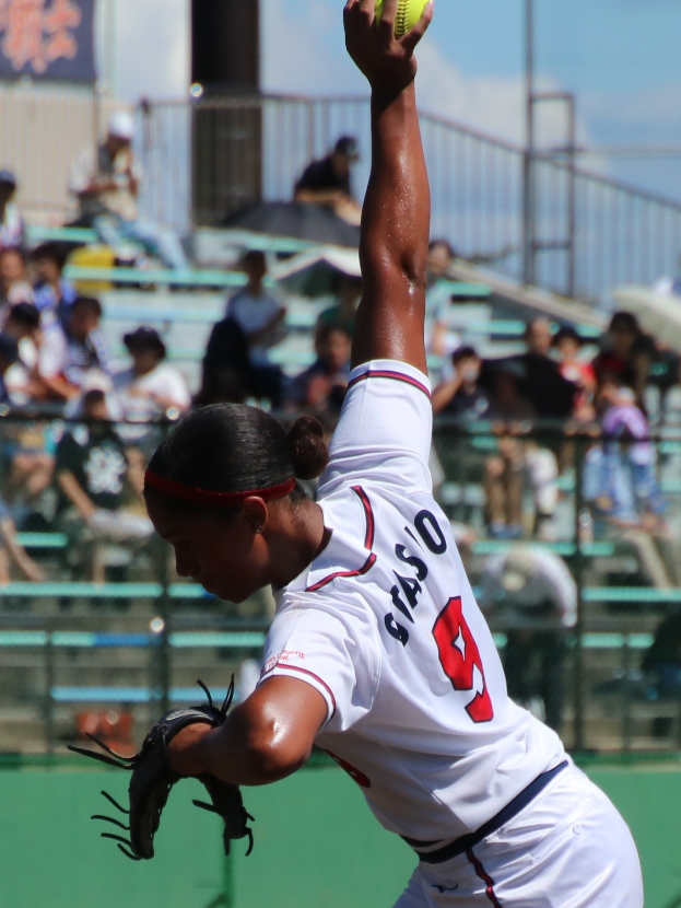 Frau in weißem Kleid und schwarzen Handschuhen wirft einen gelben Softball auf einem Feld mit Zuschauern im Hintergrund.