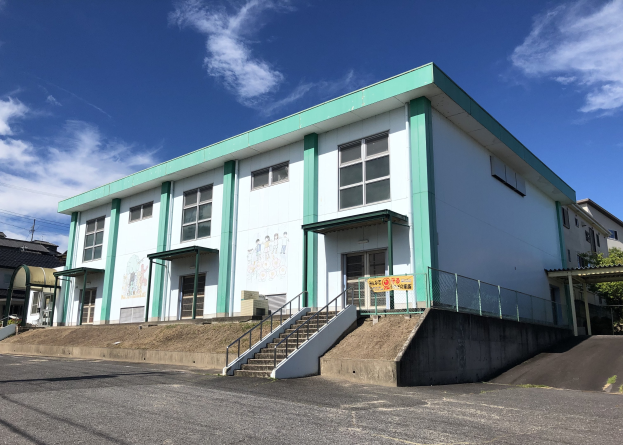 Wei├čes und grünes Geb├Ąude mit Treppe, umgeben von B├Ąumen und einer Stra├če, unter einem bew├Âlktem Himmel, als Japans erste japanische Schule identifiziert.