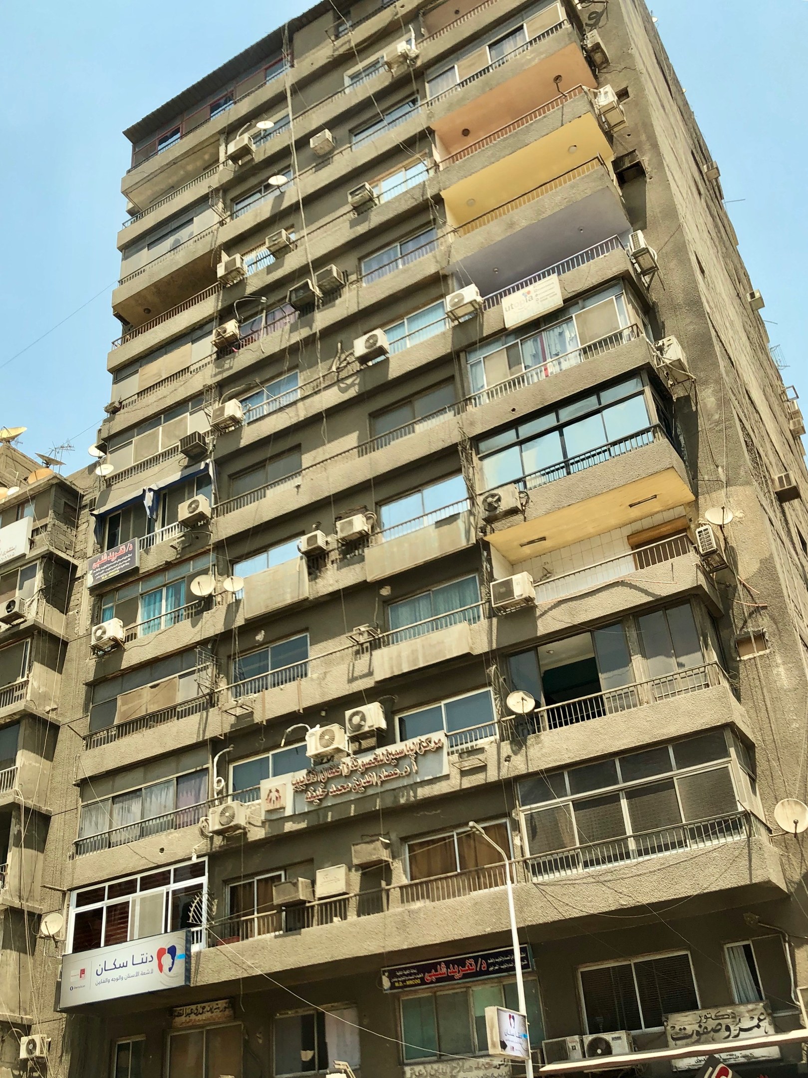Hohes Gebäude mit zahlreichen Fenstern, Balkonen, Klimaanlagen, Namensschildern, Straßenlaternen, Strommasten und Kabel gegen einen bewölkten Himmel.