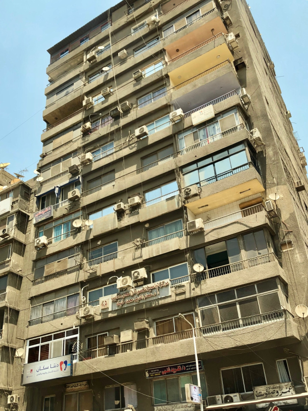 Hohes Gebäude mit zahlreichen Fenstern, Balkonen, Klimaanlagen, Namensschildern, Straßenlaternen, Strommasten und Kabel gegen einen bewölkten Himmel.