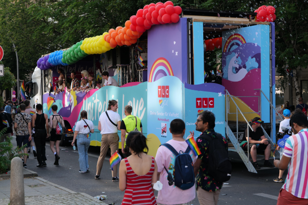Eine Gruppe von Menschen geht eine Straße entlang neben einem Lastwagen mit bunten Luftballons, einige tragen Taschen, mit Schildern an Pfählen an der Straße und Bäumen und Gebäuden im Hintergrund.