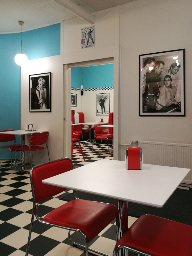 Retro-Diner-Interieur mit schwarzem und weißem Schachbrettboden, roten Stühlen und Tischen, gerahmten Bildern an der Wand, einer Deckenleuchte und verschiedenen Gegenständen auf den Tischen.