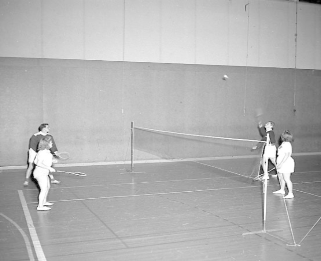 Schwarzes und weißes Bild von Menschen, die Badminton auf einem Court mit einem Netz und einer Wand im Hintergrund spielen.