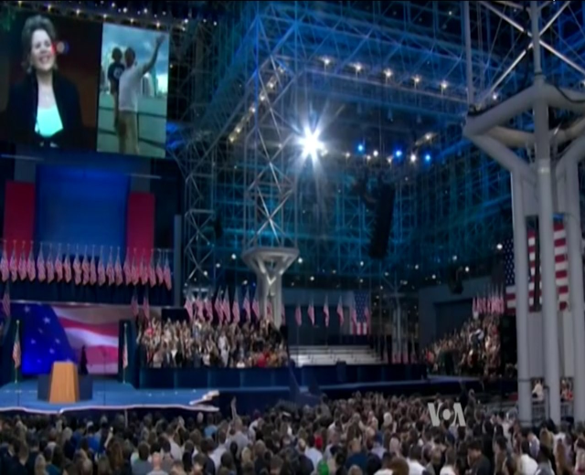 Eine große Menschenmenge sitzt vor einer Bühne mit einem Rednerpult, Mikrofon, Flaggen, einem Bildschirm und Beleuchtungsausrüstung, was auf eine Präsidenten-Debatte hindeutet.