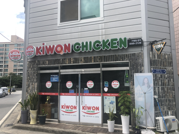 Außenansicht eines Kiwon-Chicken-Restaurants in Seoul, Südkorea, mit Glastüren und -fenstern, Topfpflanzen, einem Banner, einer Klimaanlage, einem Pfahl, Kabeln, Bäumen und Fahrzeugen auf der Straße unter einem bewölkten Himmel.