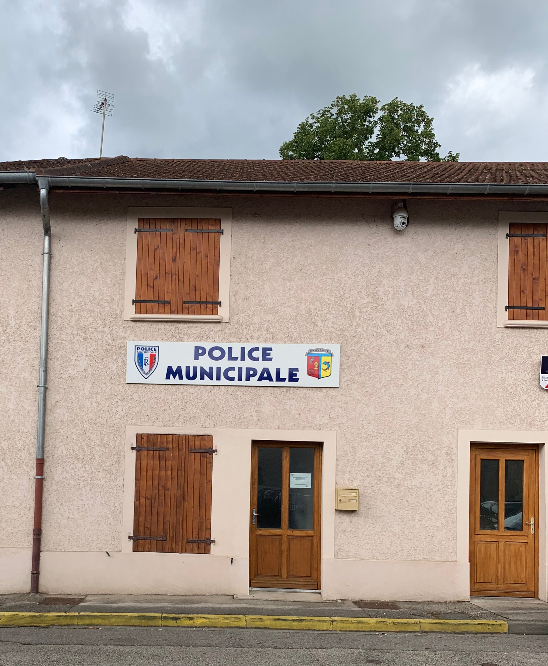 Polizei-Gebäude mit einem Schild, Fenstern, Türen, einer Namensplakette, einem Rohr, einem Baum und einem bewölkten Himmel im Hintergrund.