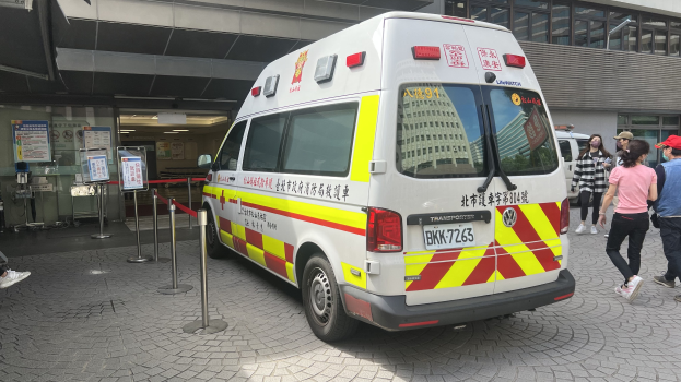 Ambulanzdienst in Hong Kong auf Schildern mit einem vor einem Gebäude geparkten Ambulanzfahrzeug, Menschen, die nach rechts gehen, und einer Person im Hintergrund.